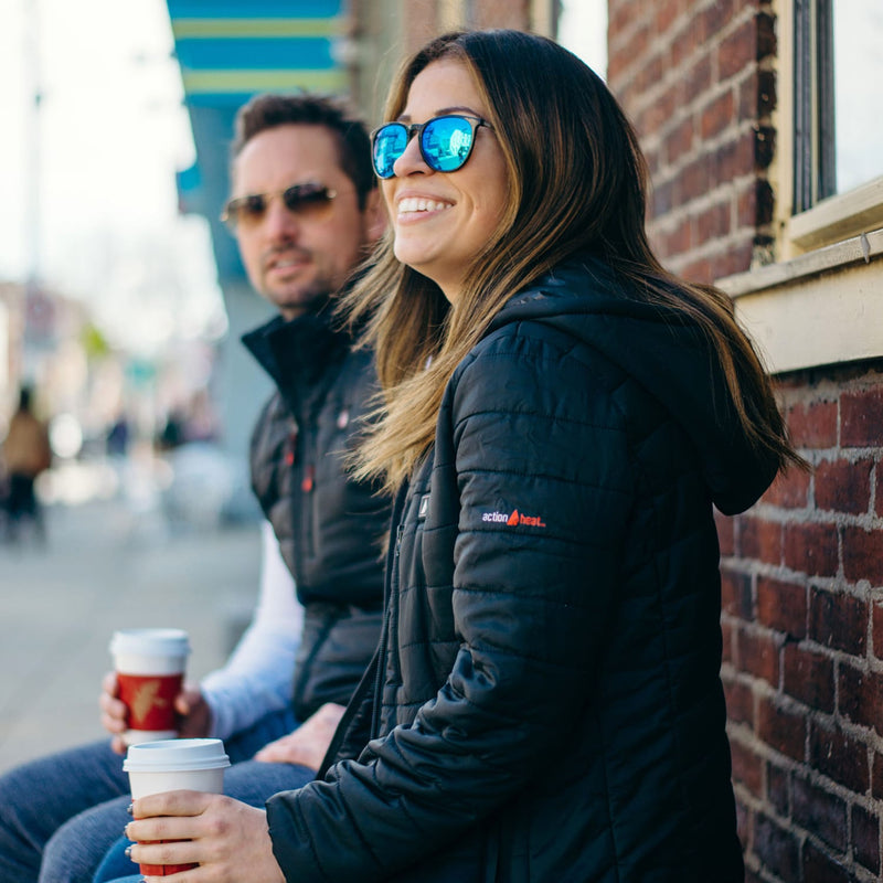 Smiling woman in black puffer jacket with blue sunglasses holds coffee, leaning against brick wall; man in similar jacket sits behind, also holding coffee.